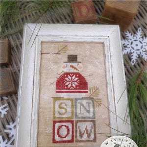 May include: A framed cross-stitch pattern titled Catching Snowflakes by Brenda Gervais, featuring a snowman wearing a red sweater with a white snowflake design, sitting atop four blocks spelling SNOW, all on a tan fabric background.