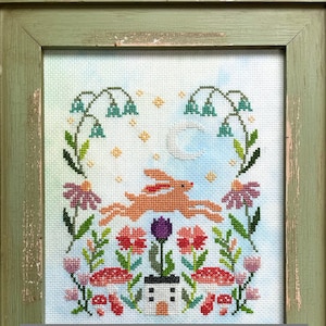 May include: A cross-stitch embroidery featuring a whimsical scene of a brown rabbit hopping through a field of flowers and mushrooms under a crescent moon. The flowers are in shades of pink, red, and purple, and the mushrooms are red with white spots. A small white house with a black door and windows sits at the bottom of the scene.
