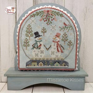 May include: A cross-stitch design of two snowmen holding hands in a winter scene with snowflakes, trees, and mistletoe. The design is framed in a blue wooden stand with the words "Mistletoe Kisses" written on the front.