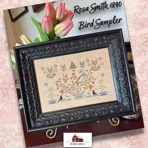 May include: Framed cross-stitch sampler with the text "Rosa Smith 1840 Bird Sampler." The sampler features a bird perched in a floral arrangement, with two small figures on either side. The frame is black with ornate detailing.