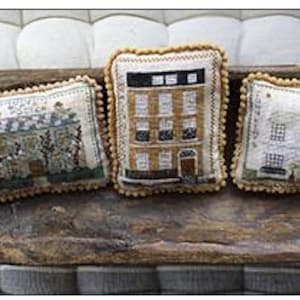 May include: Three small, square, embroidered pillow cushions with different designs. The first cushion features a green and white embroidered house with a flower garden. The second cushion features a brown and white embroidered house with a black door and windows. The third cushion features a white embroidered house with a black door and windows.