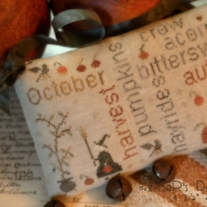 May include: A cross-stitched fabric piece with the word "October" and autumn-themed words like "harvest" and "pumpkins." The design includes a witch's hat, a broom, and a tree. The dimensions are 4 1/2" x 7 1/2".
