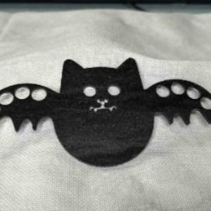 May include: A black felt bat with a cat-like face and open wings. The wings have circular cutouts. The bat is centered on a white, textured fabric.