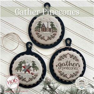May include: Three round cross-stitch ornaments with a winter theme. The ornaments feature a cabin in the woods, a red and green quilt block, and the text "gather pinecones".