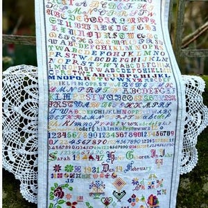 May include: A vintage cross-stitch sampler with colorful embroidered letters, numbers, and floral motifs. The sampler includes the text "Sarah 1947" and "1931 Den 14 February". The textile is hung with wooden clothespins.