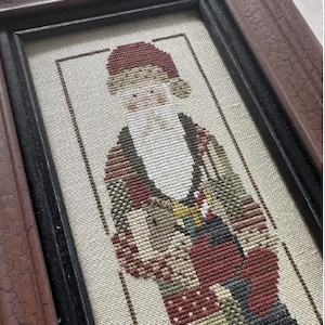 May include: A framed cross-stitch of a Santa Claus figure wearing a red hat and a patterned coat. The background is a cream-colored fabric.