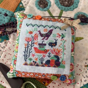May include: A square pincushion with a cross-stitched design featuring birds, a boat, and floral elements in shades of orange, green, and purple. The pincushion is trimmed with floral fabric and adorned with colorful buttons and a small bow.