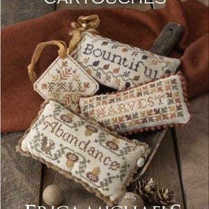 May include: An arrangement of autumn-themed needlepoint cartouches. The cartouches feature embroidered text and designs, including the words "Abundance," "Harvest," and "Bountiful." The image also includes pinecones and the text "Erica Michaels Needleart Designs."