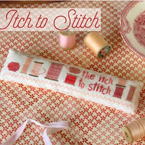 May include: A white pillow with a cross-stitch design of spools of thread and the text "The itch to stitch". The pillow is on a pink and white floral fabric with spools of thread and a pink ribbon.