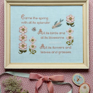 May include: Cross-stitch artwork in a light wooden frame. The piece features a light blue background with embroidered text and floral and bird motifs. The text reads "Came the spring with all its splendor..." and is surrounded by pink flowers and a blue bird.