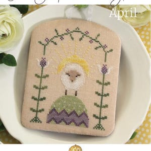 May include: A cross-stitch design featuring a sheep with a yellow halo, wearing a green and purple outfit, and the text "April". The design is framed by green vines and purple flowers. The text "The Shepherd's Year" is at the top.
