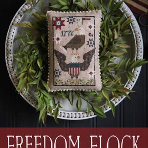May include: A beige, cross-stitched pillow with an eagle design, the year 1776, and patriotic stars and stripes. The pillow is displayed on a white beaded tray with greenery, with the text "FREEDOM FLOCK" below.
