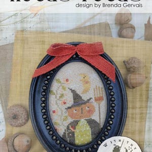 May include: An oval black frame containing a cross-stitch embroidery of a pumpkin-headed figure wearing a witch hat and cape, holding a broom. The frame is topped with a rustic orange burlap bow. Text reads Hocus Pocus, Pixie Pics, #CS382.