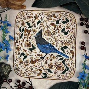 May include: A cross-stitch embroidery on light beige fabric featuring a central blue jay bird surrounded by a decorative border of pumpkins, leaves, acorns, and small animals. The design uses shades of blue, brown, green, and orange thread.