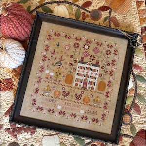 May include: A framed cross-stitch sampler on tan fabric featuring a multi-story house, a scarecrow, pumpkins, and floral borders in shades of red, brown, and cream. The design includes an alphabet, the word AUTUMN, and small animal motifs.