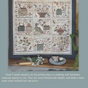 May include: A framed sampler with nine small, detailed cross-stitch panels. Each panel features a house, floral elements, and text. The sampler is displayed on a wooden surface with decorative items.