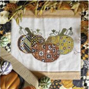 May include: A cross-stitch embroidery featuring three pumpkins in shades of orange, yellow, and blue. The pumpkins have various patterns, including stripes, checks, and polka dots, set against a white background. The embroidery is framed with a burlap border.