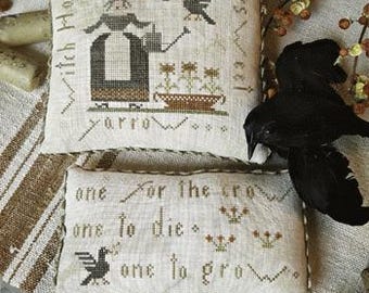 May include: Cross-stitch pattern on a white linen fabric with a witch, crows, and the text "One for the crow, one to die, one to grow." The pattern is framed with a brown fabric border.