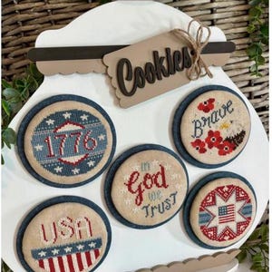 May include: A white wooden cookie jar-shaped sign with a brown lid and a tag that reads "Cookies." The sign features five round, embroidered patches with patriotic designs, including the text "1776," "Brave," "God We Trust," "USA," and a star-spangled banner.