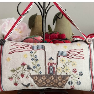 May include: A white pillow with red, white, and blue embroidered details. The pillow features a small boat with an American flag, a woman in a red dress, and a bird. The pillow is decorated with red, white, and blue ribbon.