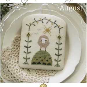 May include: A cross-stitch pattern for August from The Shepherd's Year collection. The design features a white sheep standing on a green hill with a yellow sun and green vines in the background. The pattern is on a white fabric with a brown border. The text "August" is printed in black at the top of the image.