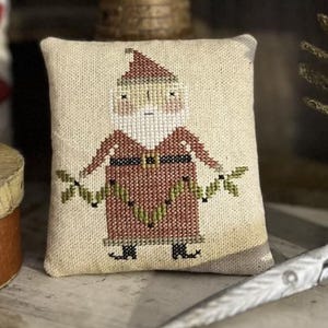 May include: A small, rectangular, cross-stitched pillow featuring a Santa Claus design. The Santa is depicted in a red robe with a matching hat, a white beard, and a green garland. The pillow has a neutral-colored background.