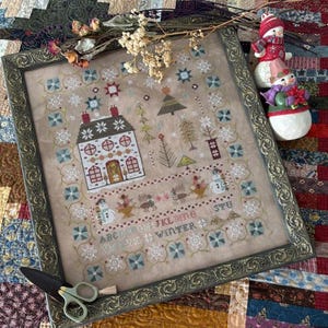 May include: Framed cross-stitch sampler with a winter theme. The design features a house, trees, snowflakes, and the word "WINTER". The frame is ornate with a dark gold finish. A pair of small scissors rests on the frame.