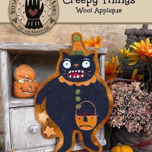 May include: A wool applique pattern titled Creepy Things by Teresa Kogut's Creative Whims. The design features a black cat wearing an orange hat and collar, holding an orange jack-o'-lantern bucket, with three green buttons on its chest.