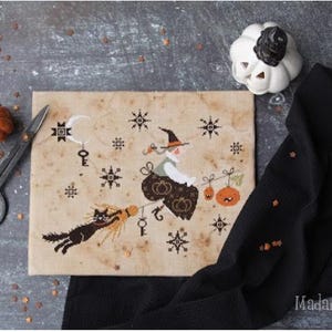 May include: A Halloween-themed cross-stitch design featuring a witch riding a broom with a black cat. The design includes pumpkins, keys, and stars. A small orange pumpkin, a white pumpkin, and black scissors are also present.