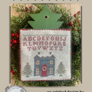 May include: A cross-stitch sampler titled "Winter Sampler" featuring a house with snow-covered roof and two pine trees. The design includes the alphabet in red thread. The sampler is surrounded by greenery and a green wooden tree.