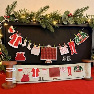 May include: A decorative display featuring a clothesline with miniature holiday-themed items including a red coat, stockings, a plaid blanket, a sweater, and a sack, suspended above a matching fabric scroll mounted on two wooden spools.