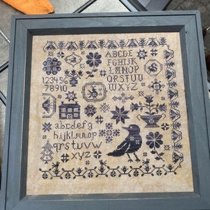 May include: A framed cross-stitch sampler with a beige background and black embroidery. The sampler features a house, a bird, flowers, and the alphabet in black thread. The text on the sampler reads: 123456 78910 ABCDE # FGHIJK LINOPS QRSTUU WXYZ * abcdefg *hijklmnop qrstuvw #xyz *