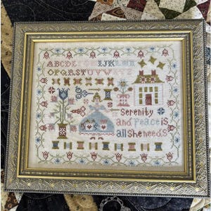 May include: Framed cross-stitch sampler with a gold-colored ornate frame. The sampler features a house, flowers, spools of thread, and the text "Serenity and Peace is all she needs". The color palette includes blue, green, red, and beige.