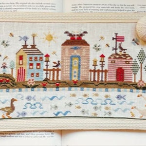 May include: A cross-stitch embroidery depicting a small town with houses, trees, and a river. The houses are colorful and have red roofs. The river is blue and has white waves. There are ducks swimming in the river. The embroidery is on a white linen fabric.