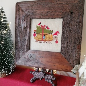 May include: A framed cross-stitch design of a turtle carrying a Christmas tree with red cardinals and ornaments. The frame is made of rustic wood and is displayed on a black metal easel.