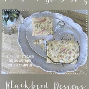 May include: Two small fabric pouches with embroidered bird designs. The pouches are on a white plate with a scalloped edge. The text "The Sweet Birds Sing" and "Blackbird Designs" are visible on the image. The image is for a cross stitch pattern.