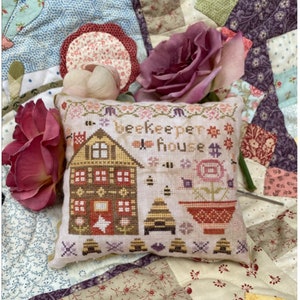 May include: A white pillow with a cross-stitch design of a beekeeper house, bees, and flowers. The text "beekeeper house" is stitched in brown thread. The pillow is on a patchwork quilt with pink and purple roses.