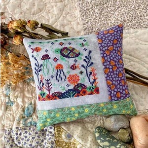 May include: A small, square decorative pillow with a cross-stitched underwater scene featuring a turtle, jellyfish, and coral in shades of orange, green, and blue. The pillow has a purple floral pattern on one side and a green floral border.