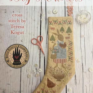 May include: A cross-stitch pattern titled "Lily's Chickens" by Teresa Kogut. The design features a rabbit holding a basket and a chicken, with floral and geometric patterns. Includes a pair of pink scissors, buttons, and a patch.