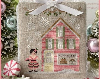 May include: A cross-stitch pattern featuring a pink and white striped building with a sign that reads "Chocolaterie". A girl in a red dress and a white hat walks towards the building in a snowy scene.