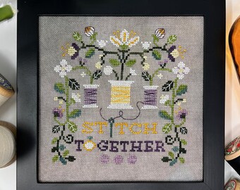 May include: A cross-stitch embroidery framed in black with a gray background. The design features two spools of thread, one purple and one yellow, with a floral arrangement above them. The text "Stitch Together" is below the spools.