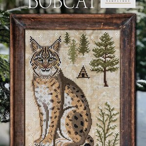 May include: Cross-stitch artwork of a bobcat, titled "The Bobcat" from "A Year in the Woods II #8." The framed piece features a detailed depiction of the animal with a forest backdrop, including trees and a small cabin. The artwork is from Cottage Garden Samplings.