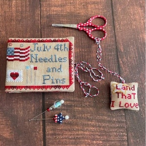 May include: A red, white, and blue needle minder with the text "July 4th Needles and Pins" and a small heart with pins. A red and white polka dot scissor is in the background. A small red, white, and blue pincushion with the text "Land That I Love" is on the right.