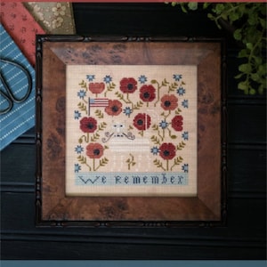 May include: Cross-stitch sampler in a wooden frame. The design features red poppies, blue flowers, and an American flag. The words "We Remember" are stitched at the bottom. The top of the image reads "Plum Street Samplers".