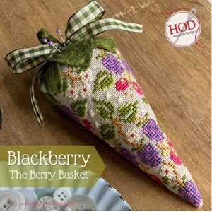 May include: A needle minder shaped like a blackberry with green leaves and purple berries. The needle minder is made of fabric and has a green felt top. It is decorated with a green and white gingham ribbon. The text "Blackberry The Berry Basket" is visible below the needle minder.