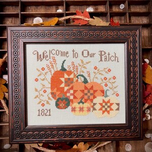 May include: A framed cross-stitch design featuring a collection of pumpkins with a patchwork quilt pattern. The text "Welcome to Our Patch" is above the pumpkins and the year "1821" is below. The frame is dark brown with a decorative border.