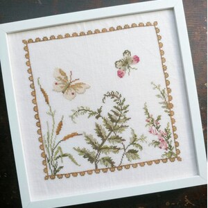 May include: A white framed cross-stitch embroidery featuring a butterfly, fern, and flowers in shades of green, brown, and pink. The embroidery is surrounded by a gold border.
