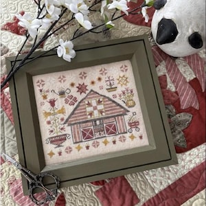 May include: A framed cross-stitch design of a red barn with a white picket fence and a flower garden. The barn is surrounded by a variety of flowers and plants, including a red wagon, a watering can, and a birdhouse. The cross-stitch is framed in a dark green frame and is sitting on a red and white quilt.