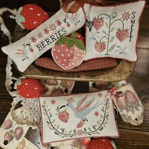May include: A collection of handmade strawberry-themed items, including a decorative pillow with the word "SWEET", a long rectangular pillow with the word "BERRIES", and a strawberry-shaped coaster. The items are in a wooden bowl.