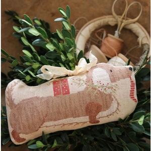 May include: A brown and white dachshund-shaped pillow with a red and white gift box, a red bow, and green holly sprigs. The pillow is made of a beige fabric and has a brown nose and eyes.
